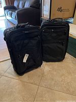 Two black softside luggage pieces standing upright on tile floor near sofa, both with handles and multiple zippered compartments.
