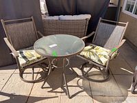 Front view of the round glass top table with two mesh fabric metal chairs with leaf patterned cushions outdoors on stone patio.