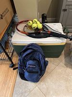 Photo showing large Igloo cooler with two tennis rackets, four tennis balls on top, and a blue JanSport backpack in front.