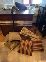 View of nine assorted throw pillows piled on the floor in front of a wooden dresser and couch.