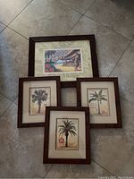 Four framed wall arts laid out together on tiled floor. One larger Hawaiian themed piece and three smaller palm tree illustrations each framed with brown wood frames.