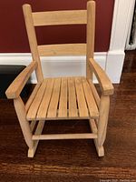 Front view of small child's wooden rocking chair with slatted seat, armrests, and rockers on a hardwood floor.