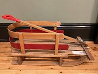 Side view of vintage style child's wooden sled showing red metal runners and wooden slats seat with red plastic handle on wooden pull rod.