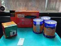 Two boxed Sony DVD players unopened, three spindles of Verbatim DVD-R discs, and a box of Compucessory CD/DVD window envelopes arranged on table.