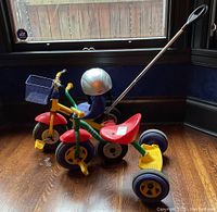 Two kids push bikes, one yellow Kettler and one green Kiddi-O, photographed indoors near window