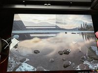 Full view of the high gloss lake photo print showing rocky shore, calm water, mountains in distance. Reflections visible on glossy surface.