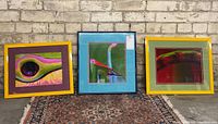 Three framed signed modern art prints displayed side-by-side on a patterned rug against a brick wall.