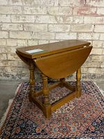 Side and angle view of a vintage solid wood small end table with drop leaves down, showing turned legs and stretcher base, set against a brick wall and on a patterned rug.