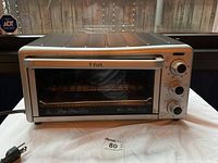 Full front view of the T-fal toaster oven showing the stainless steel exterior, glass door with horizontal handle, and three control knobs on right side.