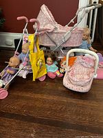 Photo showing two pink toy doll strollers, a pink doll car seat, five dolls, and a yellow toy bag arranged on wooden floor with red wall background.