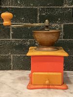 Side view of vintage manual coffee grinder showing wooden base painted red and yellow, metal bowl, and metal crank handle with yellow wooden knob