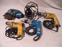 Five power tools on cloth: two yellow Dewalt drills, blue Makita screwdriver, teal Bosch jigsaw, black Porter-Cable sander.