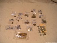 Photo of multiple foreign coins and banknotes arranged on a table, some in small plastic bags with handwritten country and date labels.