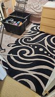 Photo showing most of the rug laid out on floor with partial room furnishings visible (dresser and storage bins). The rug has a high contrast design with swirling patterns in light on dark.