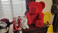 Photo showing three Marilyn Monroe Barbies on white stands and a large red foam rosette bear around 24 inches tall on top of a dark wooden dresser.