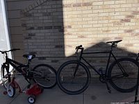 Three bicycles arranged side by side against brick wall; from left to right: red and white child's tricycle, black mountain bike, black single speed racing bike