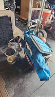 Photo of four camping lanterns, blue folding chair bag, and Igloo rolling cooler in garage floor environment.