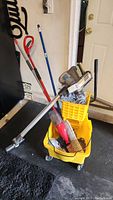 Yellow mop bucket with mop, broom, dustpan, brushes, and extension vacuum wand inside bucket at garage floor.