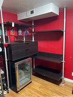 Full view of the IKEA metal tubular corner shelving system with dark wood drawers and shelves mounted near ceiling against red wall.