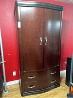 Front view of the large dark wood armoire showing the two drawers and double cabinet doors with silver handles against a red wall.