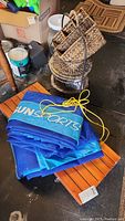 Sun Sports blue net folded on wooden slatted surface with yellow cord and black paddles visible, alongside woven basket.