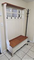 Full view of the white wooden hall tree with six black metal hooks, stained bench top, and upper shelf against cream wall.