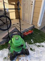 Photo showing green Lawn-Boy gas lawn mower and red manual push mower outside near porch door in snow.