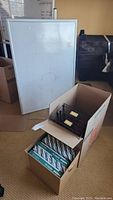 Photo showing 48x36 whiteboard standing on floor, two open cardboard boxes in front containing reams of printer paper and black metal mesh document holders.
