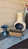 Photo showing cream chiminea, metal planter, and wicker basket on black stand against beige siding