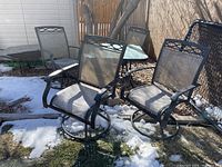 Wide view showing two swivel mesh chairs in front of square glass-top patio table missing umbrella and stand, outdoor backyard setting with grass and snow patches.