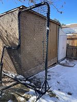 Photo of full pitching backstop showing the tall rectangular black metal frame covered with black mesh netting, set outdoors in a backyard area with some snow and grass visible.