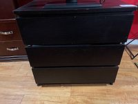 Front view of black veneered MDF three-drawer chest with visible scratches and drawer seams.