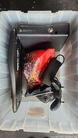 Top-down view inside plastic storage tub showing Xbox 360 console, wired controller, power adapter, and a Logitech keyboard along with a red bag of items.