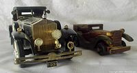 Photo showing two model cars: a black 1931 metal model car with detailed front grille and lights beside a smaller wooden model car with reddish-brown paint and gold accents.