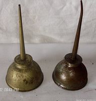 Two vintage metal oil cans with long spouts, one with a yellow brass tone and one more orange-copper tone showing patina and wear, both approximately 7 inches tall