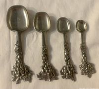 Front view of the four pewter measuring spoons arranged by size showing grape clusters on handles.