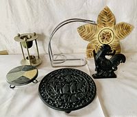 Photo of five vintage household decorative items including cast iron bird trivet, chicken napkin holder, candle holder, fruit hanger with flower base, and a small geometric patterned trivet.