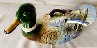 Side view of the ceramic duck soup tureen showing the green head, detailed painted feathers, removable lid with handle, and ladle placed in the opening.