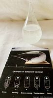 Clear drop-shaped glass barometer with white crystallized liquid inside, shown with an instructional manual featuring weather prediction symbols.