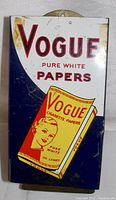 Front view showing Vogue Pure White Papers with illustrated cigarette paper pack and woman's face graphic. Minor scratches and oxidation marks visible.