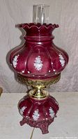 Front view of Victorian style hurricane lamp showing rose glass body with white floral decals, brass collar, and clear glass chimney with ruffled edge