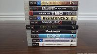 Photo showing stack of 10 PS3 game cases with visible titles on the spines arranged on a wooden surface.
