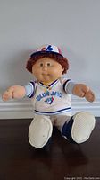 Front view of Cabbage Patch doll wearing Montreal Expos Blue Jays baseball jersey, cap, pants, and shoes, seated on a surface.