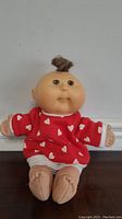 Front view of a 7-inch Cabbage Patch Kid doll with red heart-printed outfit and tuft of brown hair.