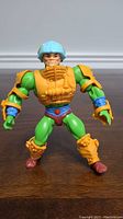Front view of green muscular Man At Arms figure with orange armor and blue helmet standing on wooden surface.