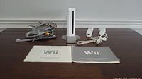 Nintendo Wii console with white base, remote controller, nunchuk, manual booklets, AV and power cables on wood surface.