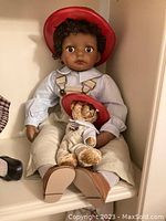 Front view of "Denzel" Little Fireman doll seated with small teddy bear doll, showing facial features, outfit details, and fireman helmet.