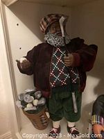 Front view of Santa Golfer doll holding a golf club and golf ball with matching golf bag filled with clubs and balls at side.