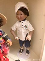 Young golfer doll holding golf ball and golf club, showing front side details with white cap and shield emblem shirt.