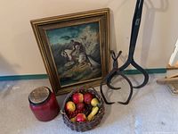 View of entire lot showing two different sized metal antique ice tongs standing upright, reproduction painting leaning against wall, willow basket filled with plastic fruit, and a red clay pot with cork top on floor.